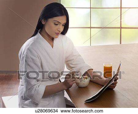 Young woman in bathrobe having breakfast while reading newspaper at dining table View Large Photo Image Picture - Young woman in bathrobe having breakfast while reading newspaper at dining table . Fotosearch