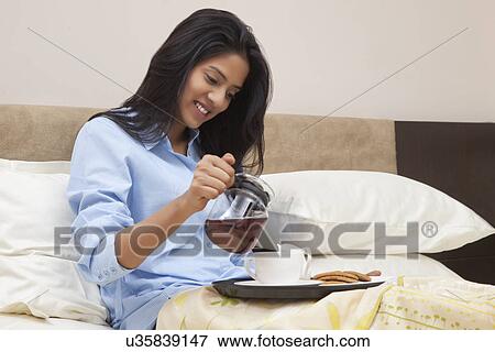 Young woman pouring tea in cup while sitting on bed View Large Photo Image Stock Photo - Young woman pouring tea in cup while sitting on bed . Fotosearch