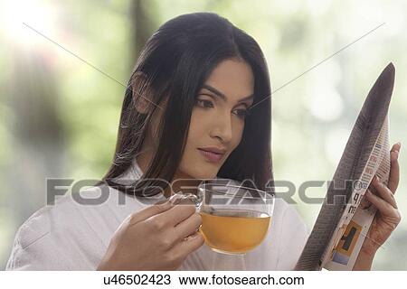 Stock Image - Young woman reading newspaper while drinking tea . Fotosearch