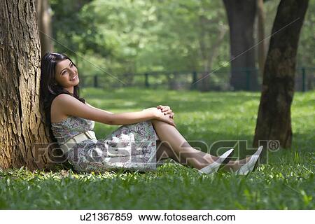 Stock Photo - Young woman smiling while leaning against a tree . Fotosearch