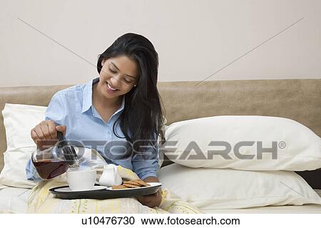 Stock Image - Young woman smiling while pouring tea in cup while sitting in bed . Fotosearch