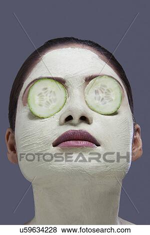 Stock Photo - Young woman with facial mask and cucumber over her eyes . Fotosearch