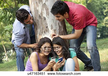 Young women sharing messages while men peeking from behind in lawn View Large Photo Image Stock Image - Young women sharing messages while men peeking from behind in lawn . Fotosearch