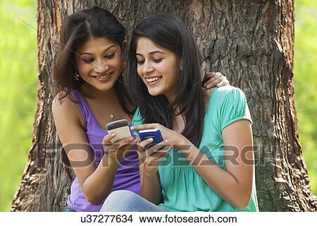 Picture - Young women sitting against tree trunk using cell phone . Fotosearch