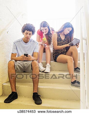Stock Image - Group of friends (14-15) sitting on steps and using digital tablet and cell phones. Fotosearch