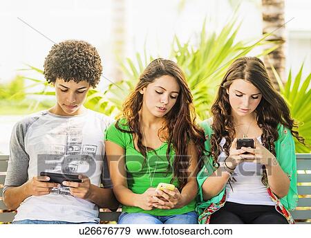 Stock Photo - Group of friends (14-15) sitting on bench and texting. Fotosearch