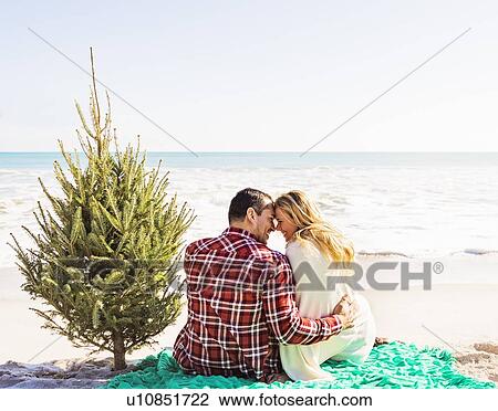 Stock Image - Loving couple sitting on beach with tree. Fotosearch