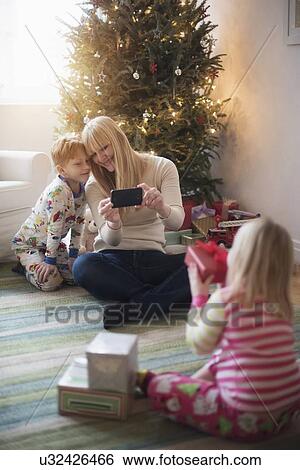 Stock Photograph - Mother with children (4-5, 6-7) opening christmas presents and taking photos. Fotosearch