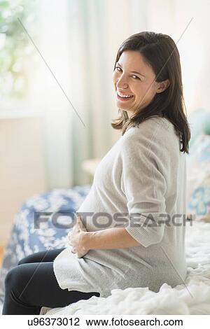 Stock Image - Pregnant woman holding ultrasound, sitting on bed smiling. Fotosearch