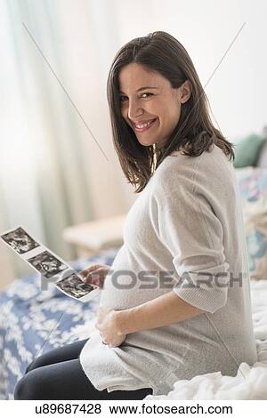 Stock Photo - Pregnant woman holding ultrasound, sitting on bed smiling. Fotosearch