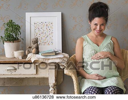 Stock Photo - Pregnant woman sitting in chair touching abdomen smiling. Fotosearch