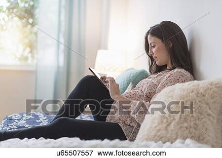 Pregnant woman sitting on bed using tablet View Large Photo Image Stock Photo - Pregnant woman sitting on bed using tablet. Fotosearch