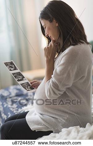 Stock Photo - Pregnant woman watching ultrasound, sitting on bed. Fotosearch