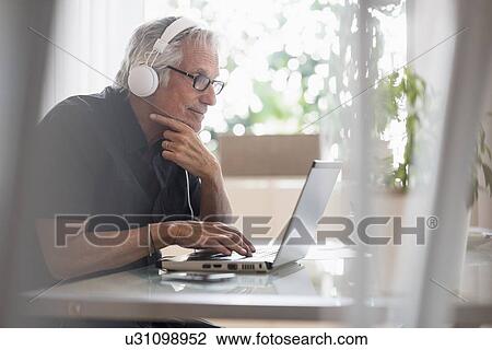 Senior man sitting in home office wearing headphones and using laptop View Large Photo Image Stock Image - Senior man sitting in home office wearing headphones and using laptop. Fotosearch