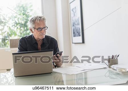 Senior man sitting in home office, using laptop and cell phone View Large Photo Image Stock Image - Senior man sitting in home office, using laptop and cell phone. Fotosearch