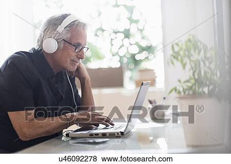 Senior man sitting in home office wearing headphones and using laptop View Large Photo Image Stock Photo - Senior man sitting in home office wearing headphones and using laptop. Fotosearch