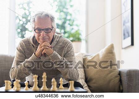 Stock Photograph - Senior man sitting in living room and playing chess. Fotosearch