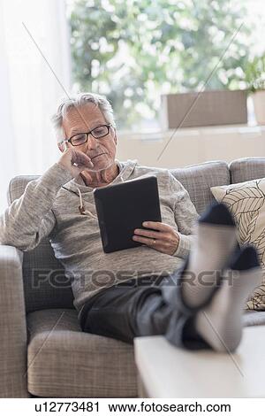 Stock Image - Senior man sitting on couch in living room and using tablet pc. Fotosearch