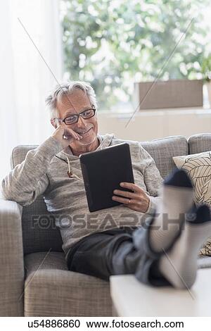 Senior man sitting on couch in living room, using tablet pc and smiling View Large Photo Image Stock Image - Senior man sitting on couch in living room, using tablet pc and smiling. Fotosearch