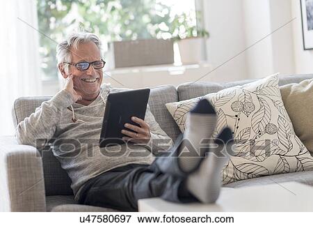 Stock Photo - Senior man sitting on couch in living room, using tablet pc and smiling. Fotosearch