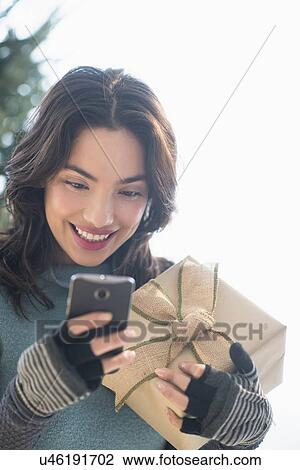 Smiling young woman holding gift and text messaging View Large Photo Image Stock Image - Smiling young woman holding gift and text messaging. Fotosearch