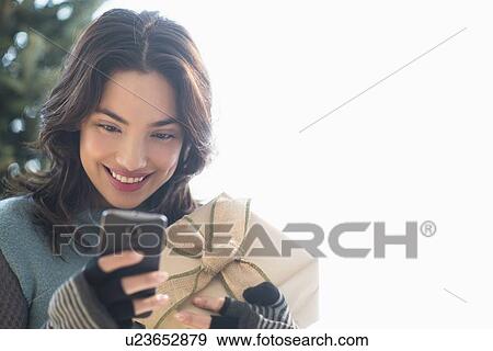 Smiling young woman holding gift and text messaging View Large Photo Image Stock Photo - Smiling young woman holding gift and text messaging. Fotosearch