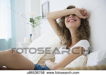 Smiling young woman sitting on bed and covering face with arm View Large Photo Image Stock Photo - Smiling young woman sitting on bed and covering face with arm. Fotosearch
