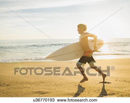 Stock Image - Teenage boy (14-15) with surfboard running on beach. Fotosearch