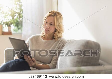 Stock Image - Woman sitting on sofa and using tablet pc. Fotosearch