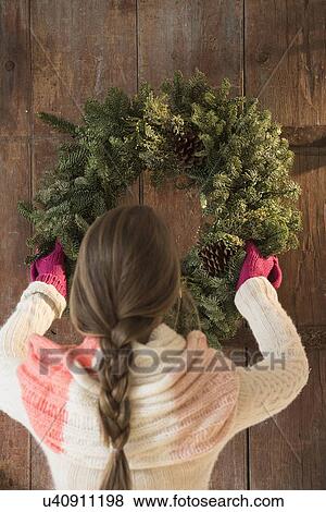 Stock Photo - Young woman hanging christmas wreath on door. Fotosearch