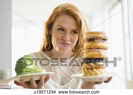 Picture - Young woman holding plate with donut and with broccoli. Fotosearch