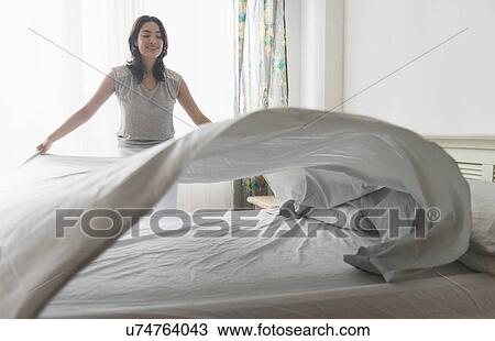 Stock Image - Young woman spreading sheet on bed. Fotosearch