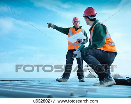 Stock Photo - Building foreman holding plans and directing apprentice on factory roof. Fotosearch