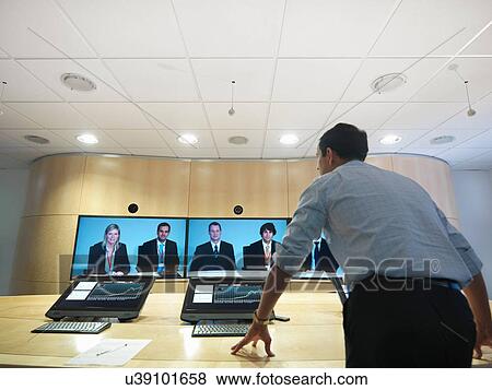 Businessman standing in meeting room addressing colleagues on video conference screens View Large Photo Image Stock Photo - Businessman standing in meeting room addressing colleagues on video conference screens. Fotosearch