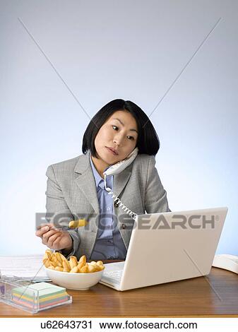 Stock Image - Businesswoman eating and working at desk. Fotosearch