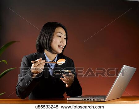 Stock Photography - Businesswoman eating and working at desk. Fotosearch