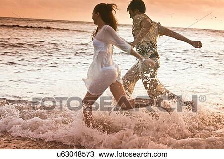 Stock Image - Couple running in waves at beach. Fotosearch