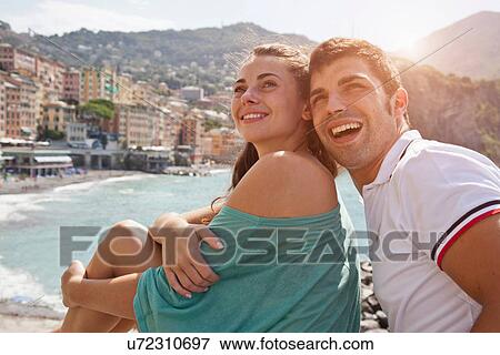 Stock Photo - Couple sitting on stone wall by ocean. Fotosearch