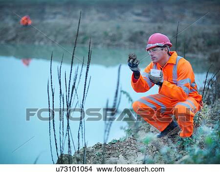 Picture - Ecologist taking soil sample from bank next to water on quarry site. Fotosearch