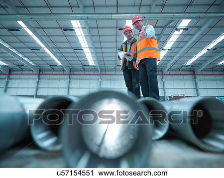Stock Image - Foreman and apprentice checking construction pipes in partly completed factory. Fotosearch
