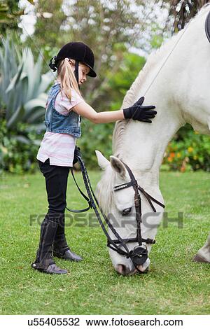 Stock Image - Girl petting grazing horse in park. Fotosearch