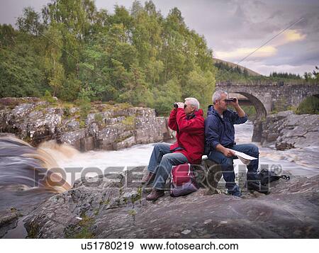 Stock Photo - Hikers sitting by flowing Scottish river looking through binoculars. Fotosearch