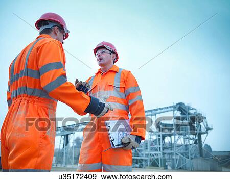Stock Photo - Low angle view of quarryman advising apprentice in front of stockpile quarry machinery. Fotosearch