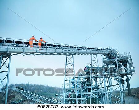 Stock Image - Low angle view of workmen climbing steps of screening conveyor at quarry. Fotosearch