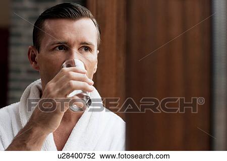 Stock Image - Man in bathrobe drinking water by window. Fotosearch