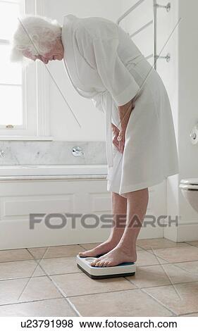 Stock Photo - Older woman weighing herself in bathroom. Fotosearch