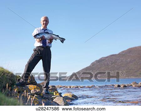 Stock Photograph - Portrait of businessman holding up freshly caught salmon by loch at fish farm. Fotosearch