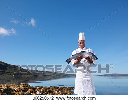 Stock Image - Portrait of chef holding freshly caught salmon by loch. Fotosearch