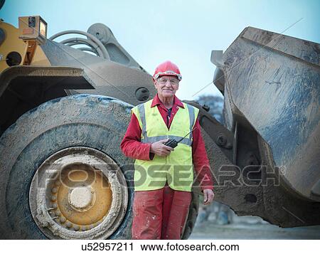 Stock Image - Portrait of workman standing in front of digger on quarry site. Fotosearch