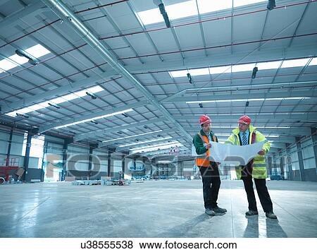 Stock Photo - Site foreman advising apprentice over plans in partly completed factory. Fotosearch
